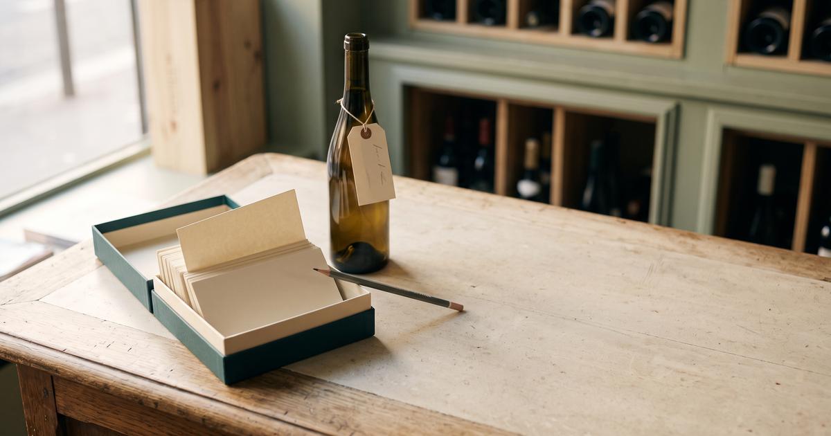 Comptoir en bois clair d'une cave à vin, une boîte à fiches vert sapin ouverte sur des cartes cartonnées vierges, un crayon posé en travers, et une bouteille portant une étiquette de papier nouée au goulot, devant des rangements à bouteilles vert sauge