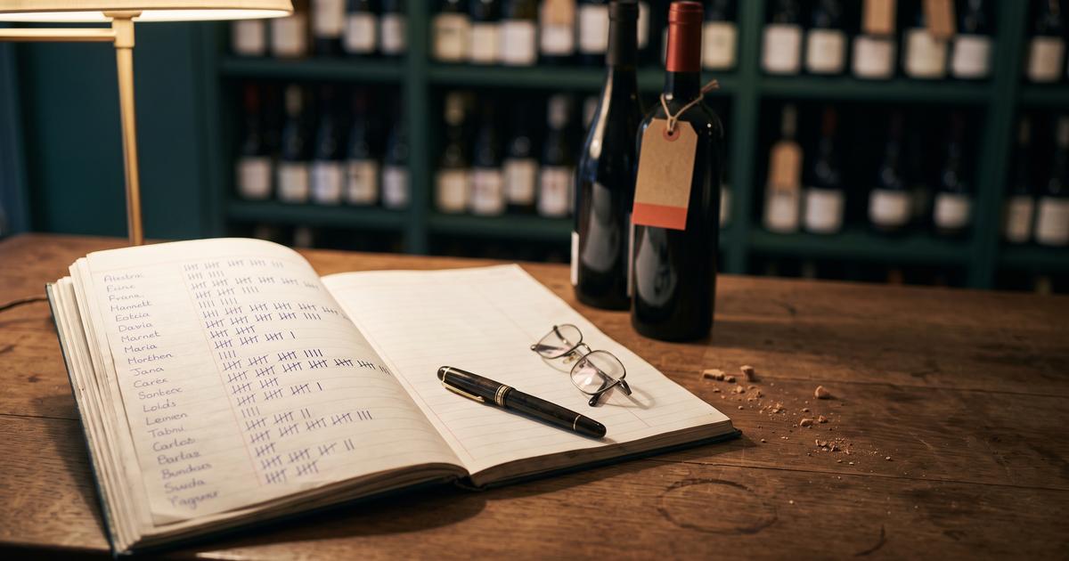 Comptoir d'une cave à vin en fin de journée : un carnet ouvert sur une liste de prénoms manuscrits, chacun suivi d'une rangée de bâtons de comptage d'espacement inégal, un stylo plume posé en travers de la page et deux bouteilles devant un rayonnage vert sombre